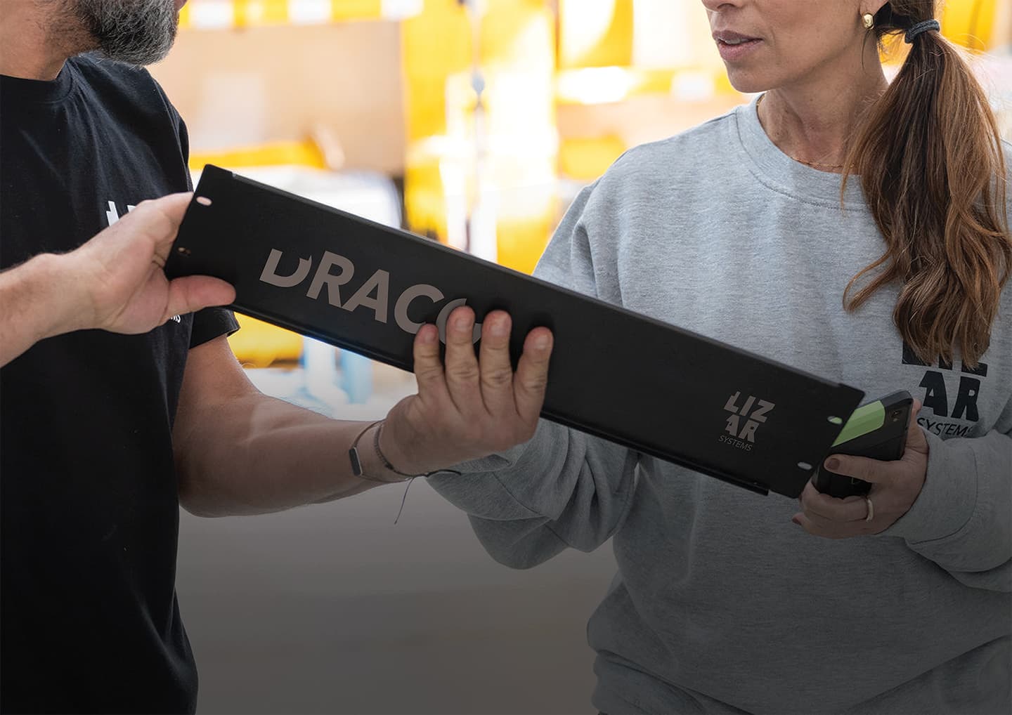 Two people in LIZAR Systems clothing handing over a long black DRACO-branded module in a production area
