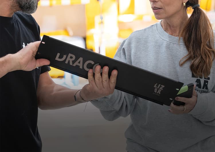Two people in LIZAR Systems clothing handing over a long black DRACO-branded module in a production area
